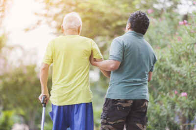 younger man helps older man walk with can outside
