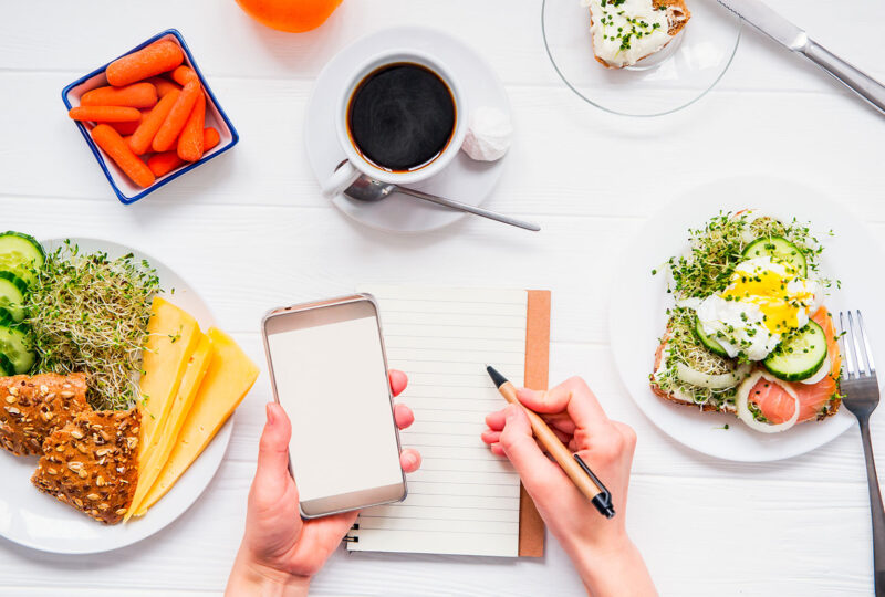 hands writing in a notebook and holding a phone on a table of food