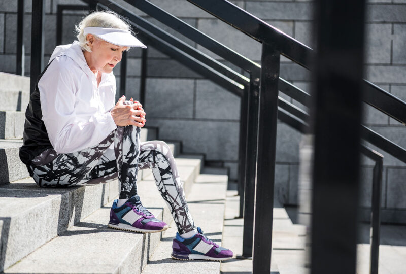 older woman sitting outside on steps in workout clothes holding knee