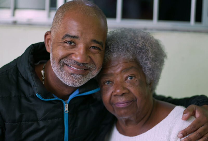 man with arm around older woman, both smiling at camera