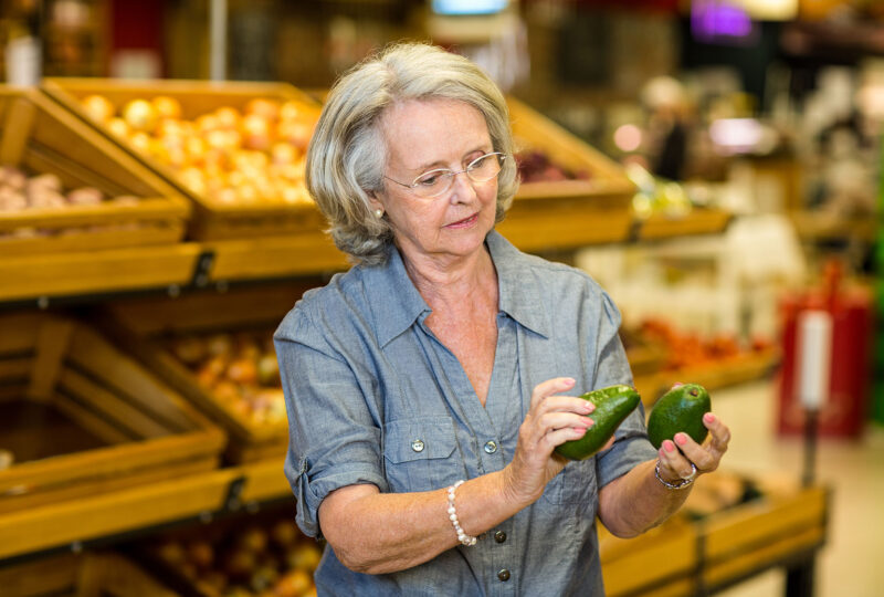older woman in grocery store holding two avocados