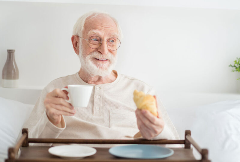 man in bed holding croissant and mug over tray