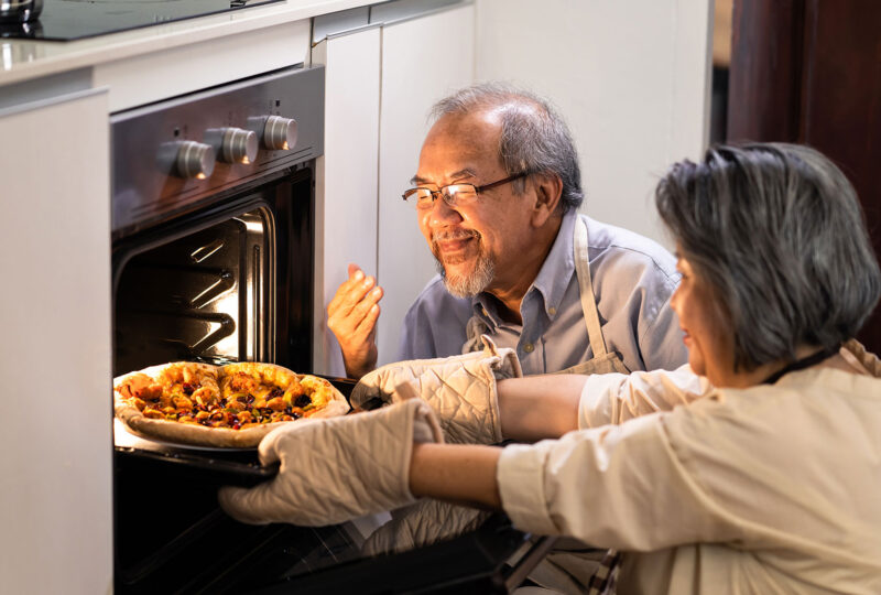 older woman pulling pie out of oven while man smells it