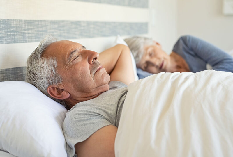 older man sleeping in bed with wife in background sleeping
