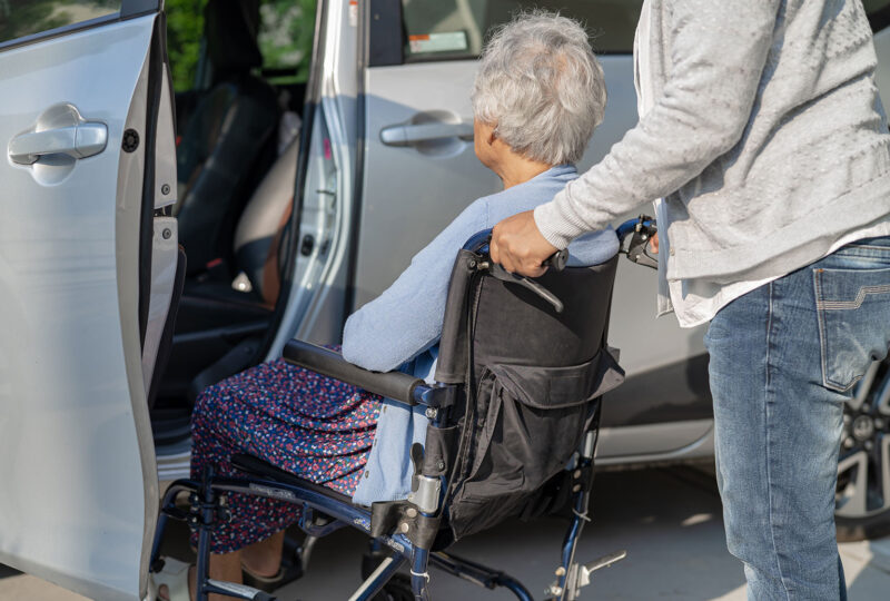 person helping senior woman in wheelchair into car