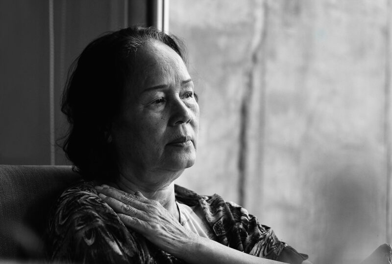 older woman sitting near a window with a hand on her chest