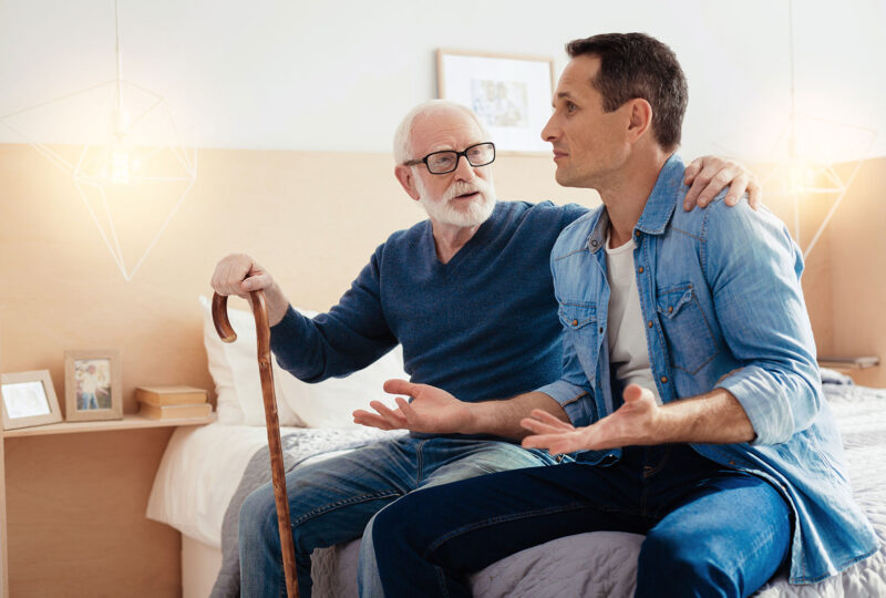 young man having animated conversation with older man holding can as they both sit on the edge of a bed
