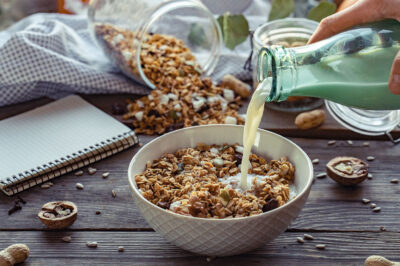 Female hand holding glass bottle pouring milk in cereal granola flakes bowl with nuts seeds raisins on brown wooden table background