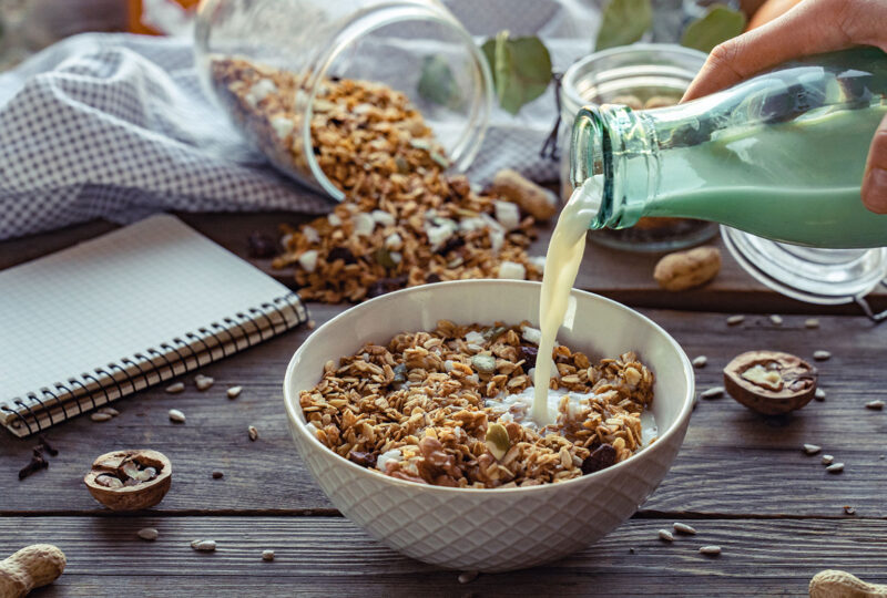 Female hand holding glass bottle pouring milk in cereal granola flakes bowl with nuts seeds raisins on brown wooden table background