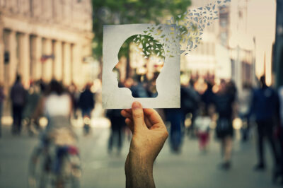 Hand holding a paper sheet with human head icon broken into pieces over a crowded street background. Concept of memory loss and dementia disease. Alzheimer's losing brain and memory function.
