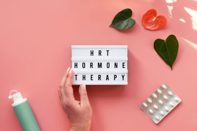 Text HRT Replacement Therapy on light box in hand. Menopause, hormone therapy concept. Pink background with exotic leaves, flowers, pills, estrogen gel.