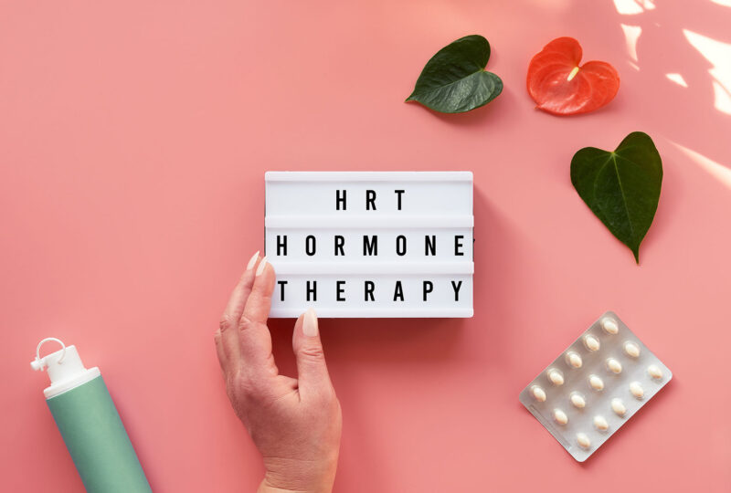 Text HRT Replacement Therapy on light box in hand. Menopause, hormone therapy concept. Pink background with exotic leaves, flowers, pills, estrogen gel.