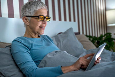 Mature woman wearing blue light blocking glasses with amber lenses, lying in bed before sleep, looking at tablet screen