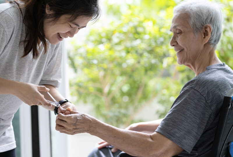 younger woman clipping nails of older woman in wheelchair, both smiling